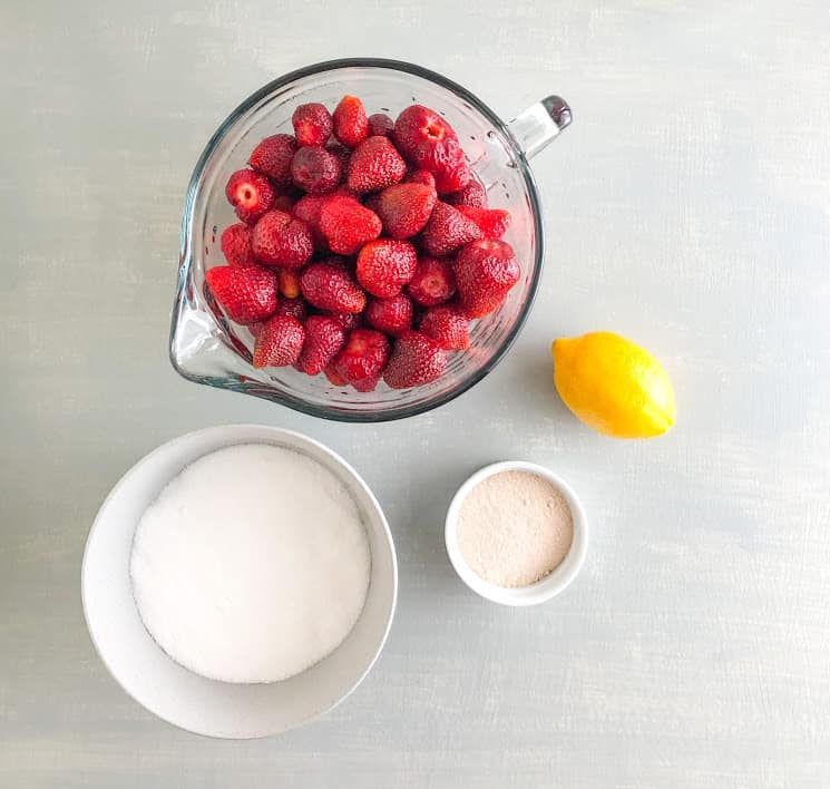 Whole strawberries in large glass measuring bowl, one lemon, round gray dish with sugar, small white round ramekin of pectin