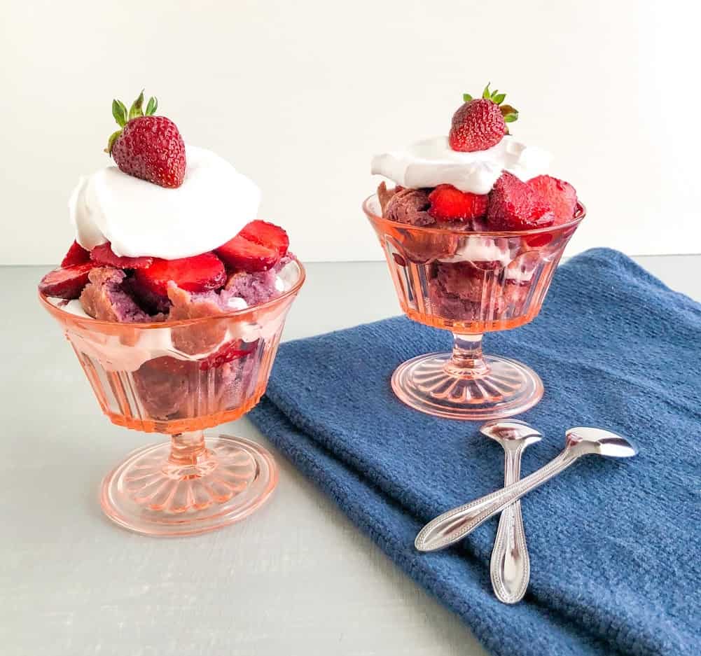 Mini strawberry shortcake trifles with purple sweet potato biscuits in pink vintage footed dessert glasses with sliced strawberries, cool whip, biscuits and whole strawberry on top of cool whip, blue dish towel, small silver teaspoons