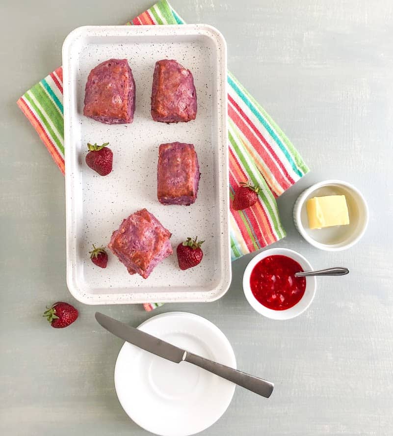 Purple sweet potato biscuits and whole strawberries on white sheet pan with gray splatters on multi-colored dish towel, small white round plates with silver butter knife, small round white bowl with strawberry lemon freezer jam and small silver spoon, small ramekin with butter