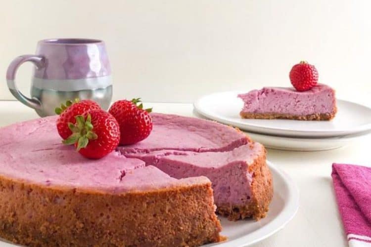 Whole purple sweet potato cheesecake with one slice on round white plate topped with whole strawberries, next to pink and white kitchen towel, two small white plates stacked in background with one slice of cheesecake topped with a whole strawberry, purple and gray coffee mug in background