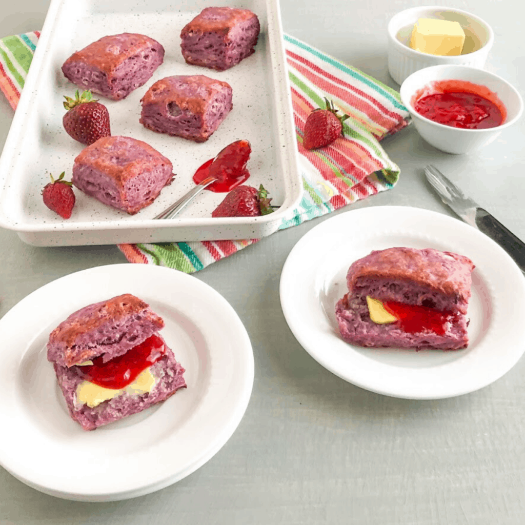 Purple sweet potato biscuits on white sheet pan with while strawberries and small silver spoon with strawberry lemon freezer jam, small white round plate with split purple sweet potato biscuit with pat of butter and strawberry lemon freezer jam, small round white bowl with strawberry lemon freezer jam, small round white ramekin with butter, silver butter knife, striped multi-color kitchen towel
