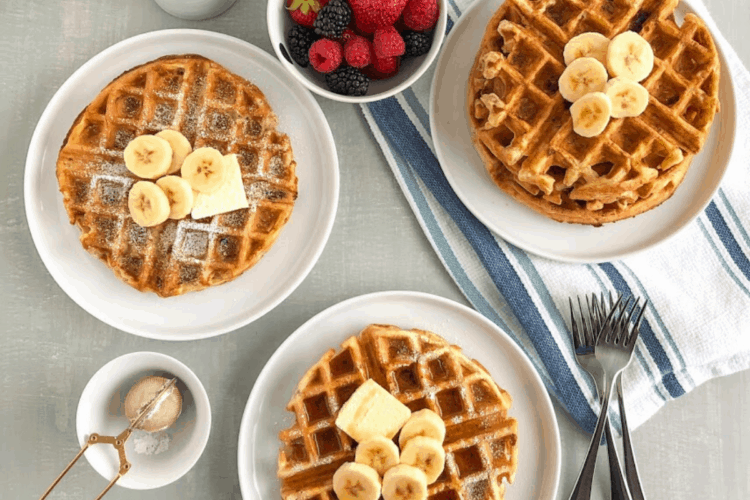 Simple banana oatmeal waffle on small white round plate with pat of butter, sliced bananas powdered sugar, small pitcher pouring maple syrup on waffles, small round white bowl with black stripes with mixed berries, stack of waffle on small white round plate in background on blue stripped kitchen towel with silver forks