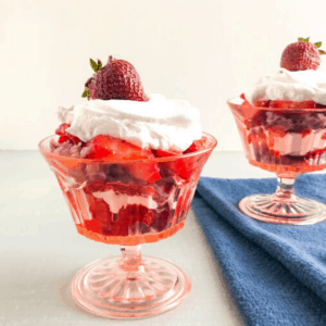 Strawberry shortcake trifles with purple sweet potato biscuits in pink vintage footed dessert glasses with sliced strawberries, cool whip, biscuits and whole strawberry on top of cool whip, blue dish towel