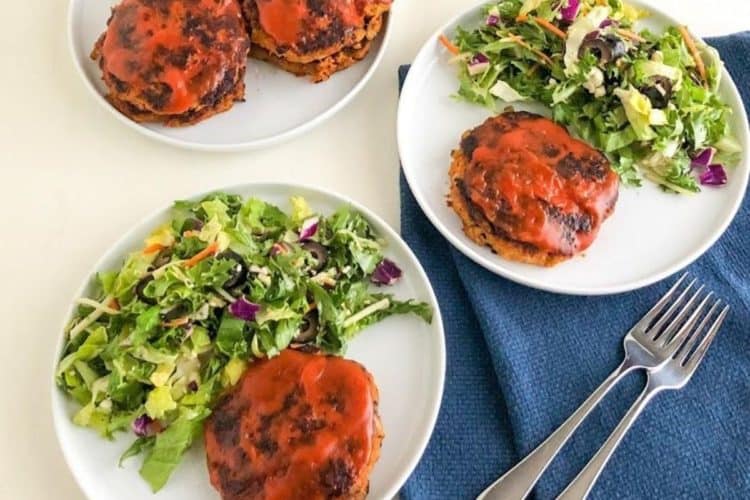 Two small round white plates with Turkey veggie meatloaf patty and green salad, one plate on dark blue kitchen towel and two silver forks with third small round white plate with additional meatloaf patties