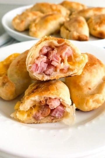 Baked ham and cheese empanadas on small white round plate with one empanada cut in half to show the inside. additional empanadas in the background on a round white plate. on white kitchen towel with blue and gray stripes.