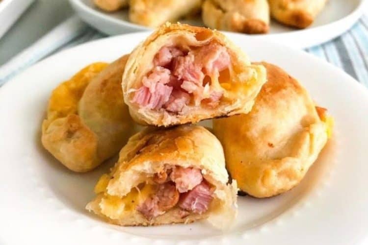 Baked ham and cheese empanadas on small white round plate with one empanada cut in half to show the inside. additional empanadas in the background on a round white plate. on white kitchen towel with blue and gray stripes.