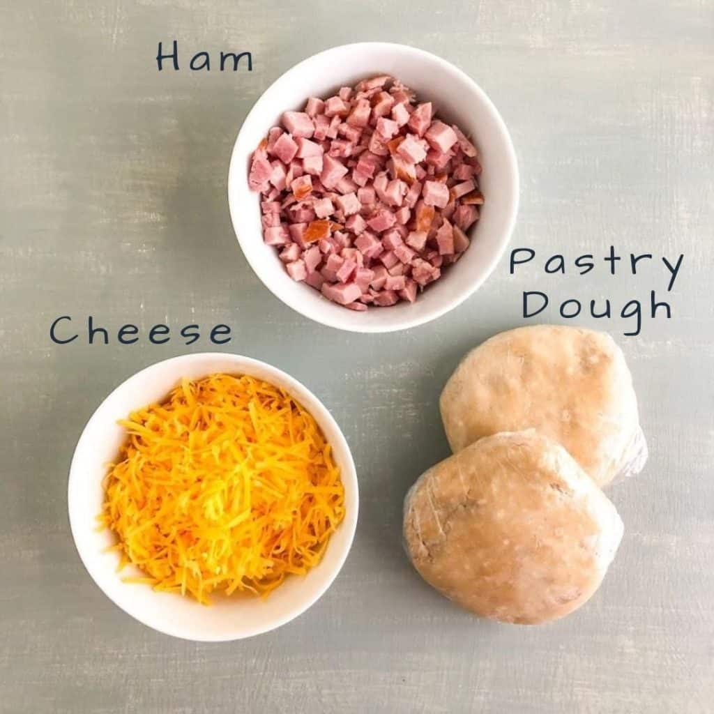 Diced ham and shredded cheese in separate round white bowls, two discs of pastry dough wrapped in plastic wrap