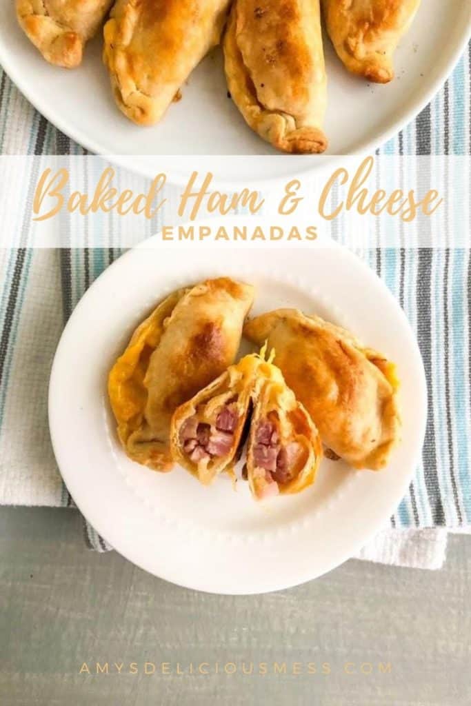 Baked ham and cheese empanadas on small white round plate with one empanada cut in half to show the inside, additional empanadas in the background on a round white plate on white kitchen towel with blue and gray stripes.