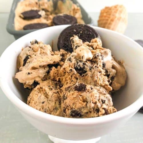 Scoops of coffee Oreo ice cream in round white footed dessert bowl with whole Oreo. Loaf pan with ice cream and waffle cones in the background.