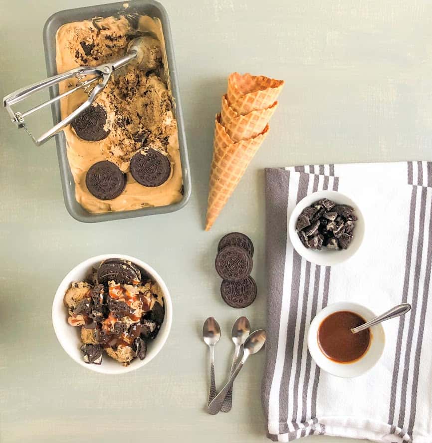 Ice cream sundae in round white bowl, ice cream in loaf pan topped with whole Oreo's and ice cream scoop in pan as well.  Small silver spoons next to a stack of Oreo's next to waffle cones.  White kitchen towel with gray stripes with small round white bowls of crushed Oreo's and salted caramel sauce with small silver spoon on top.