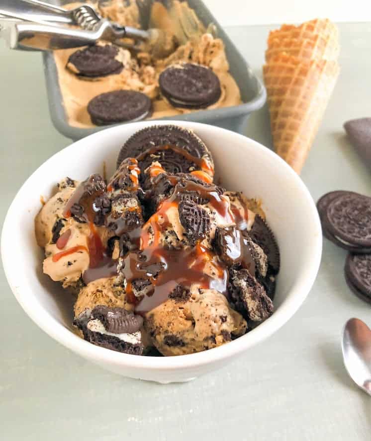 Scoops of  ice cream in round white footed dessert bowl with whole and crushed Oreo's and salted caramel sauce. Loaf pan with ice cream and waffle cones in the background next to waffle cones, Oreo's and small silver spoon.