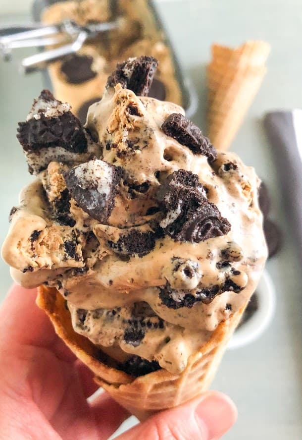 Hand holding waffle cone with ice cream topped with Oreo chunks.  Loaf pan with extra ice cream and ice cream scoop, plus waffle cones in the background