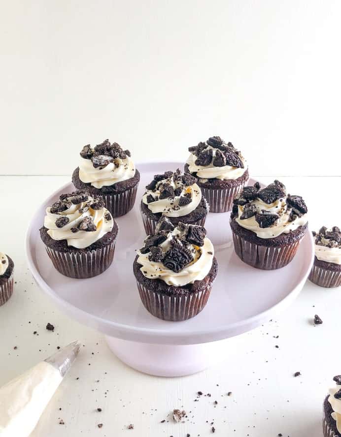 Oreo cupcakes with cream cheese buttercream on light pink cake stand with piping bag of buttercream underneath, additional cupcakes and Oreo crumbs on the ground