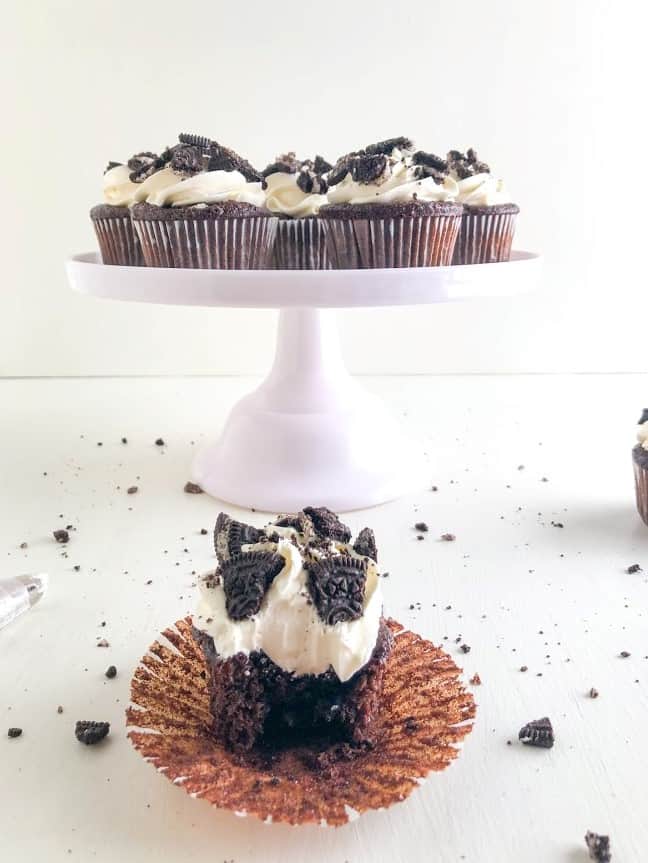 Oreo Cupcakes with Cream Cheese Buttercream with liner peeled back, extra cupcakes in the background on light pink cake stand, Oreo crumbs sprinkled the ground