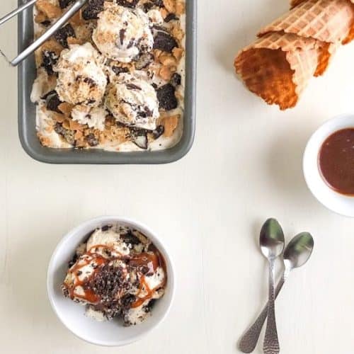No churn ice cream in small round white bowl with salted caramel sauce, loaf pan with ice cream and ice cream scoops on top, small silver spoons, waffle cones, and small round white bowl of salted caramel sauce on the side.
