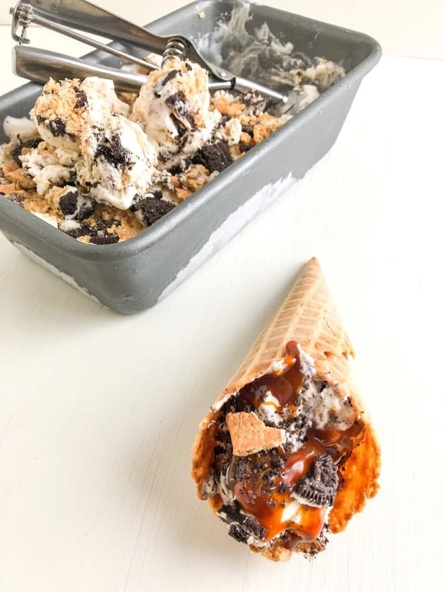 Waffle cone with ice cream topped with salted caramel sauce laying on its side, loaf pan with remaining ice cream and ice cream scoops and cookie scoop in the background.