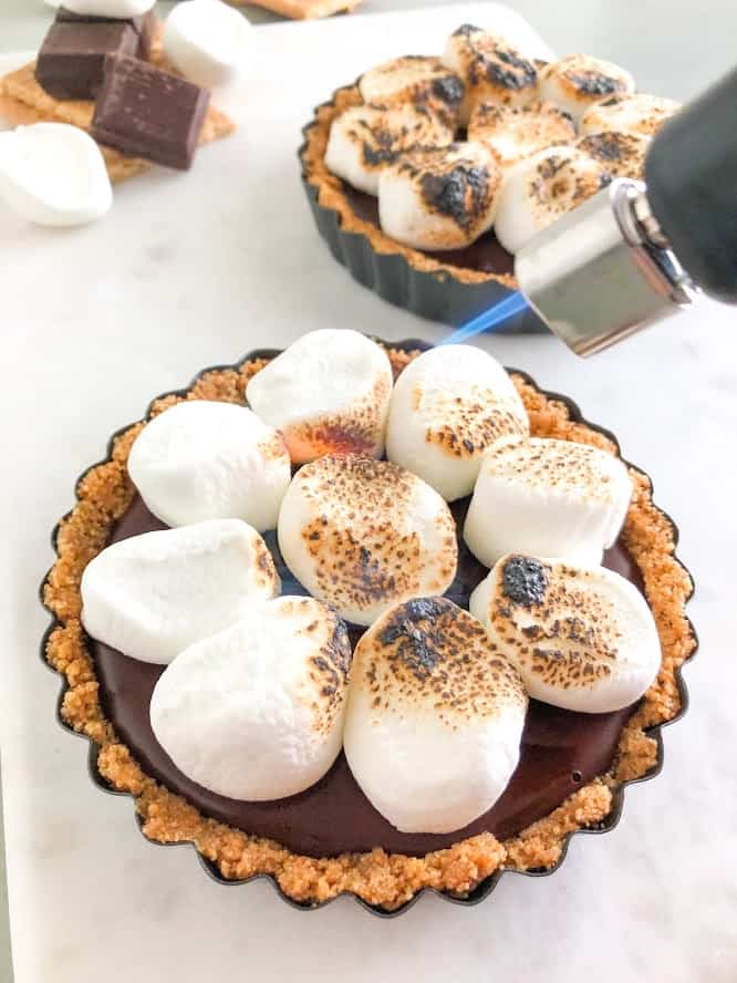 Using a kitchen torch to toast marshmallows on s'mores tart on marble board with second s'mores tart with toasted marshmallows, additional chocolate squares, marshmallows, and graham crackers in the background