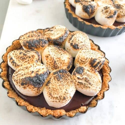 S'mores tart on marble board with additional s'mores tart, marshmallow, graham crackers, and squares of chocolate in the background
