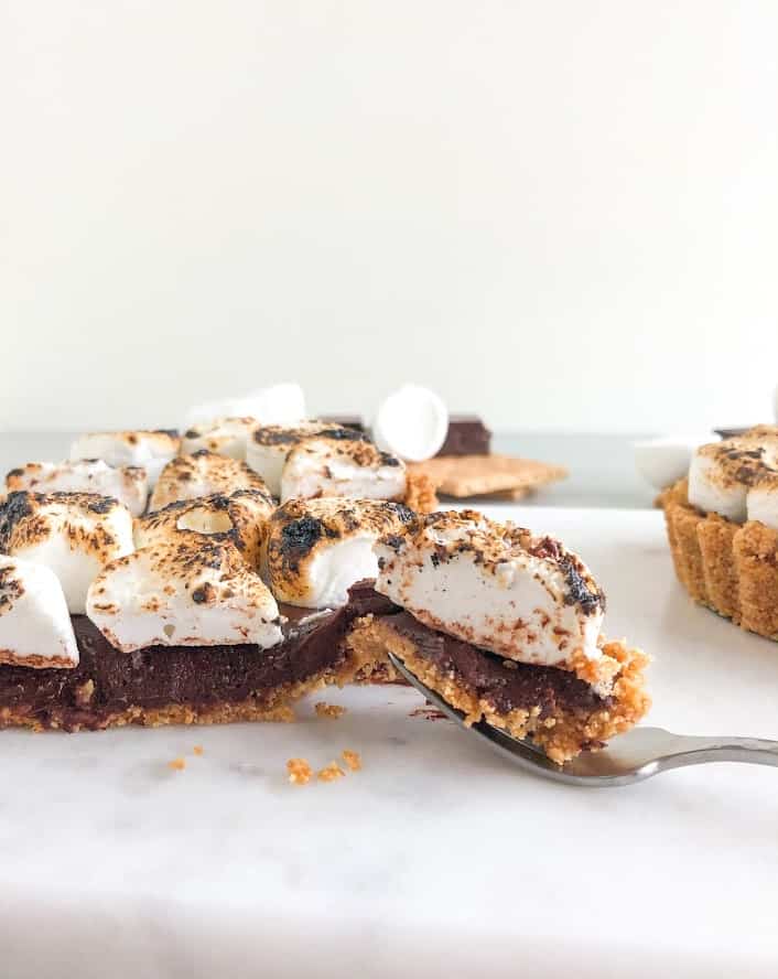 S'mores tart cut in half with a bite on small silver fork on a marble board with another s'mores tart next to it and additional chocolate, marshmallows, and graham crackers in the background