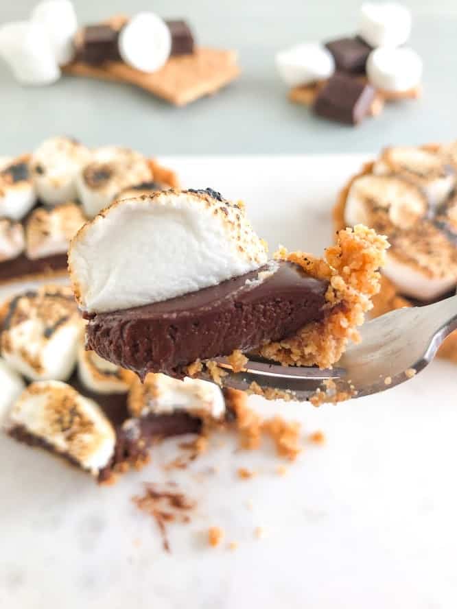 Silver fork with a bite on s'mores tart and s'mores tarts, marshmallows, chocolate, and graham crackers in the background