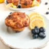 Lemon blueberry muffin on small round light blue plate with lemon slices and blueberries, round white medium plate with additional muffins, lemon slices, and blueberries. Whole lemon and blueberries on the background for decoration