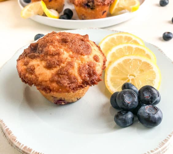 Lemon blueberry muffin on small round light blue plate with lemon slices and blueberries, round white medium plate with additional muffins, lemon slices, and blueberries. Whole lemon and blueberries on the background for decoration