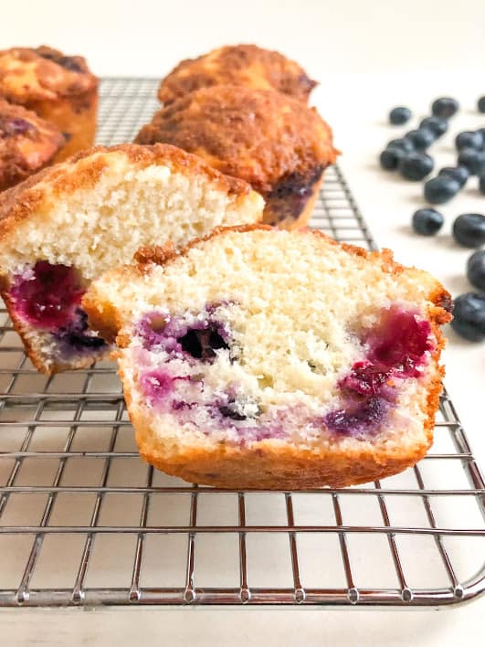 Lemon blueberry muffin cut in half on silver metal cooling rack with additional muffins and whole blueberries in the backgound