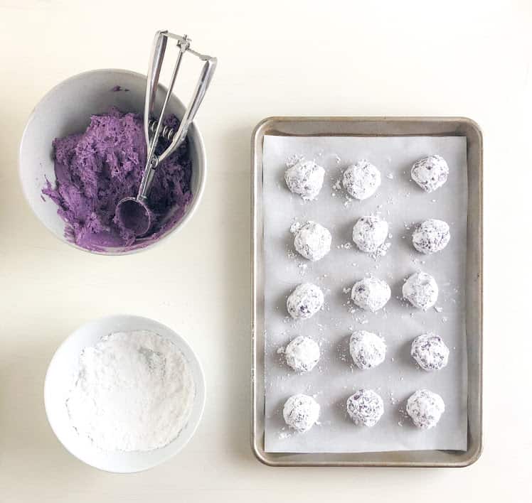 cookie dough balls rolling in powdered sugar and on parchment lined baking sheet, bowls of powdered sugar and cookie dough with cookie scoop.