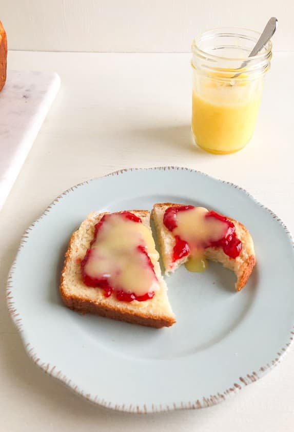 Slice of bread on small round light blue plate topped with butter, strawberry lemon freezer jam, and lemon curd.  Jar of lemon curd with small silver spoon in the background