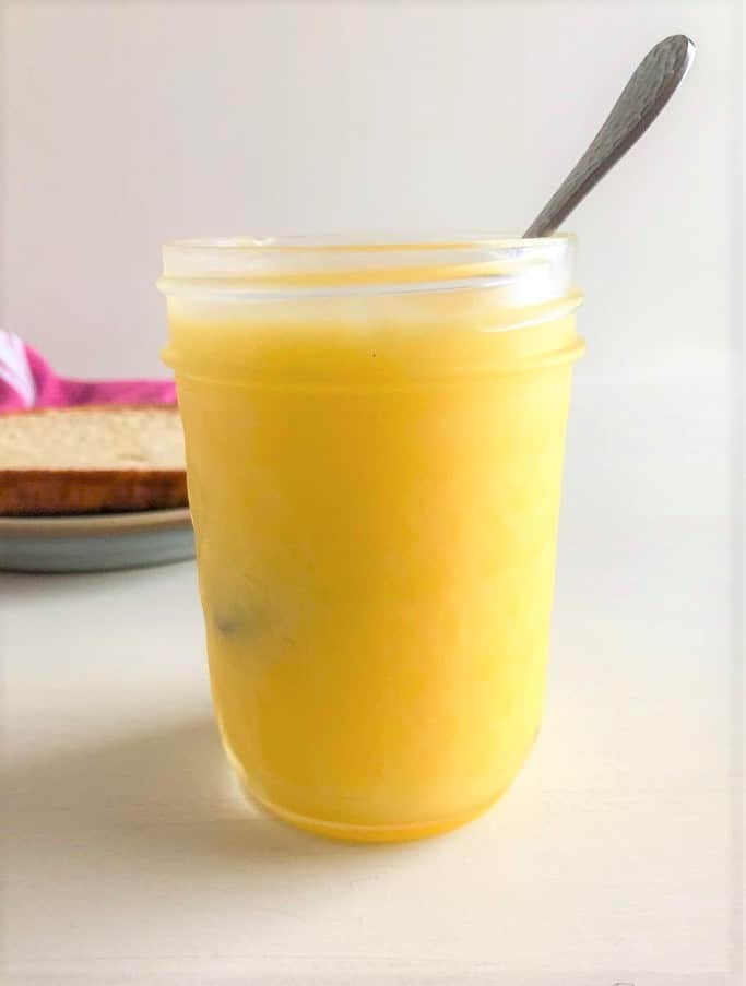 Mason jar with lemon curd, small silver spoon in jar.  Slice of bread on small round light blue plate and pink and white striped kitchen towel in the background