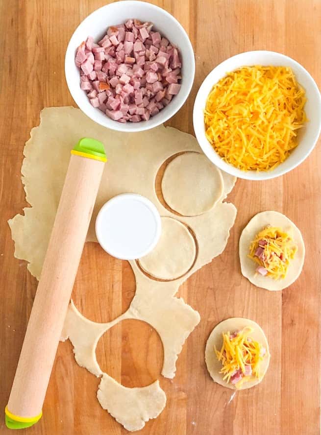 Diced ham and shredded cheese in separate round white bowls, pasty dough rolled out with Joseph Joseph rolling pin on top.  Circles of pastry dough cut out, ham and cheese filling on two circles of dough.  Everything on a wooden cutting board.