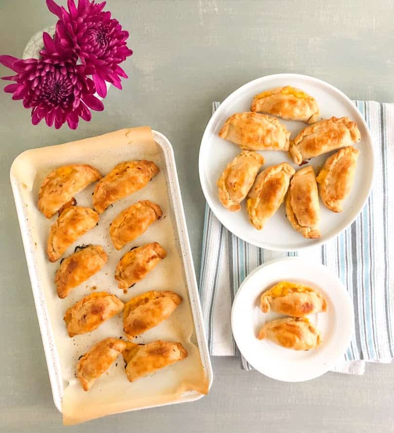 Baked ham and cheese empanadas on small white round plate, additional empanadas on a round white plate on white kitchen towel with blue and gray stripes. Parchment lined white baking pan with baked empanadas. Small vase with gray cracks and purple flowers.