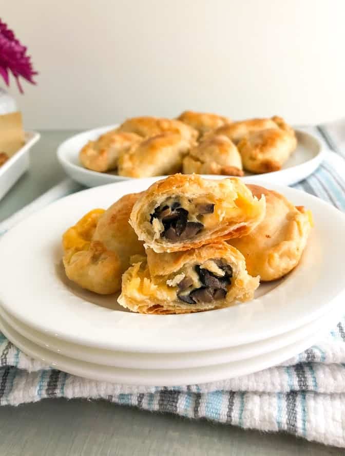 Baked black olive and cheese empanadas on small white round plate with one empanada cut in half to show the inside. additional empanadas in the background on a round white plate on white kitchen towel with blue and gray stripes. Small vase with gray cracks and purple flower in the background