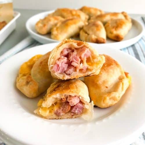 Baked ham and cheese empanadas on small white round plate with one empanada cut in half to show the inside. additional empanadas in the background on a round white plate. on white kitchen towel with blue and gray stripes. Small vase with gray cracks and purple flower in the background