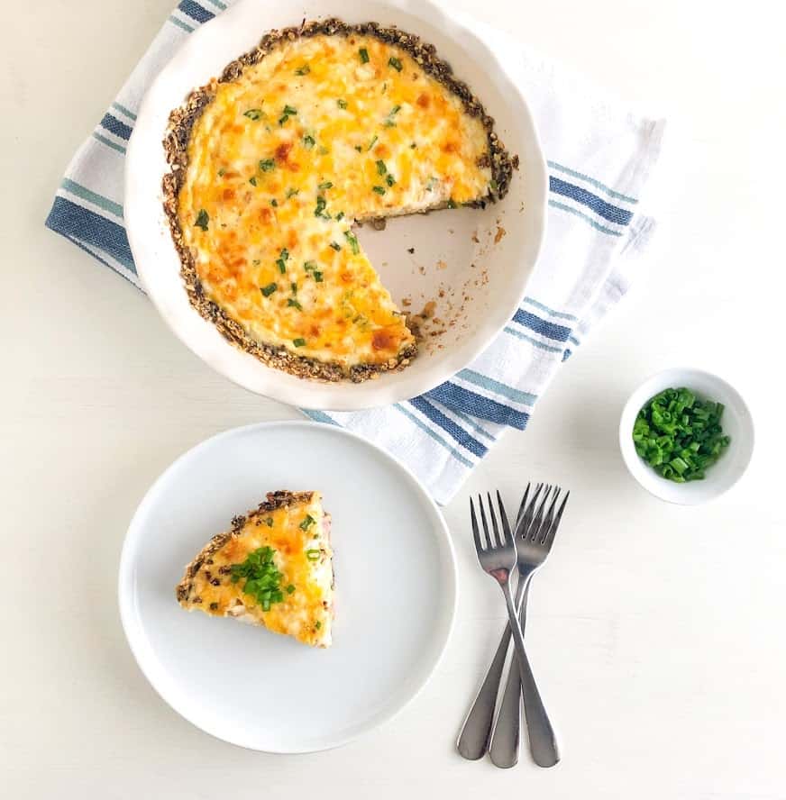 Slice of quiche on small round plate topped with sliced green onions, quiche in Emile Henry round white pie dish on white towel with blue stripes, silver forks and small round bowl of sliced green onions
