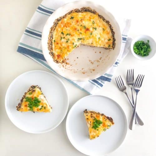 Slices of mushroom crust quiche on small white round plates topped with sliced green onions, next to three silver forks, emile henry flour pie dish with remaining quiche on white kitchen towel with blue stripes, small white round bowl with sliced green onions