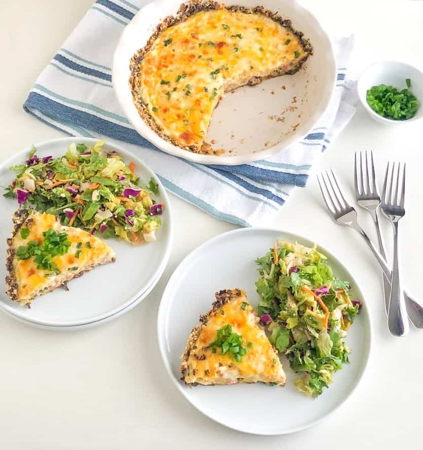Slices of quiche on small white round plates topped with sliced green onions and side green salad, next to three silver forks, Emile Henry round pie dish with remaining quiche on white kitchen towel with blue stripes, small white round bowl with sliced green onions