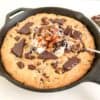 Skillet cookie in black cast iron pan with two silver spoons, scoops of ice cream, and salted caramel sauce. Part of chocolate bar with dried fruit and nuts in the background.