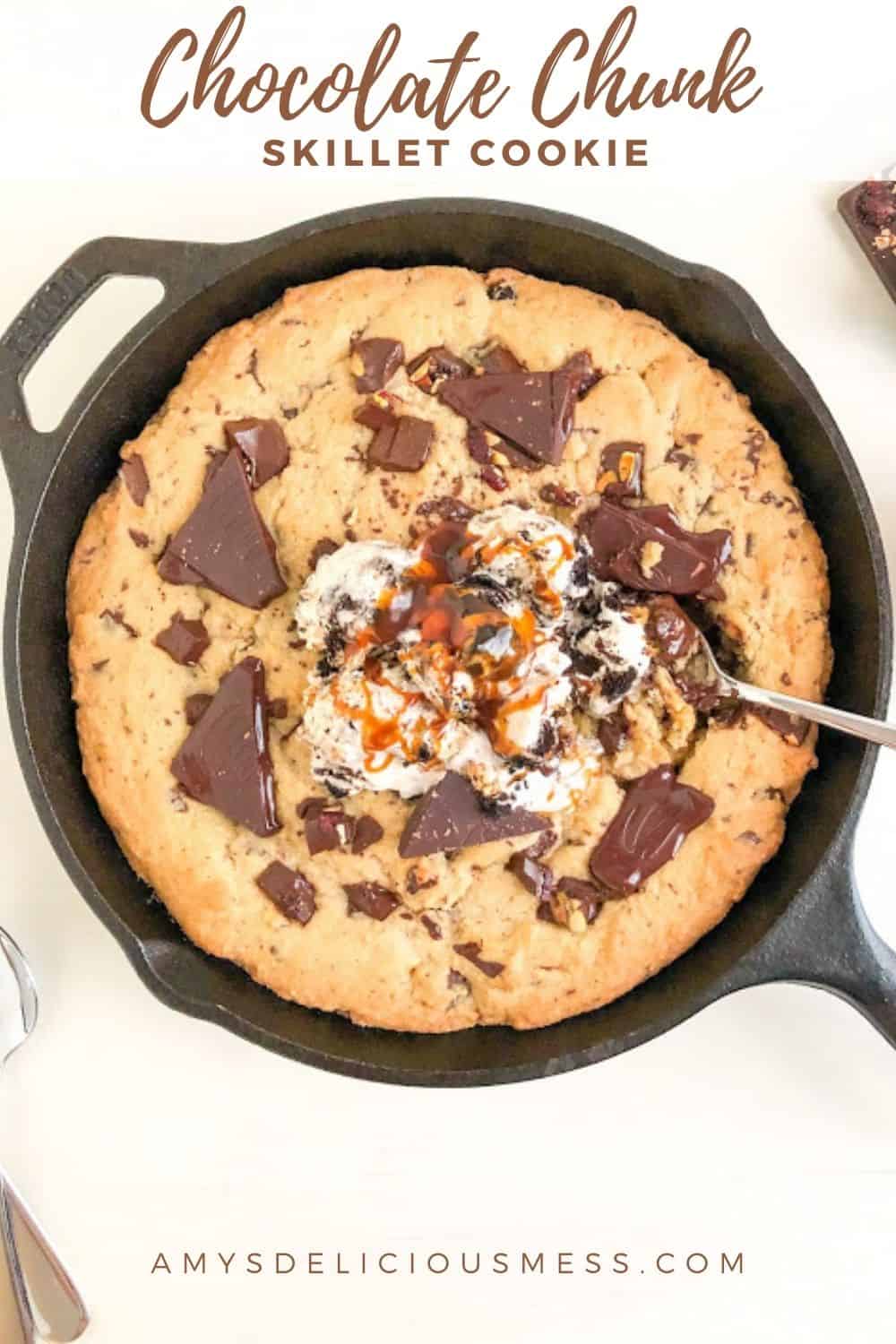 Skillet cookie in black cast iron pan with silver spoons, scoops of ice cream, and salted caramel sauce. Part of chocolate bar with dried fruit and nuts in the background. Small silver spoons next to the cast iron skillet.