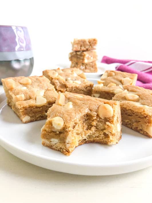 blondies on round white plate with on missing a bite. Purple and gray coffee cup, stack of blondies, and pink kitchen towel with white stripes in the background