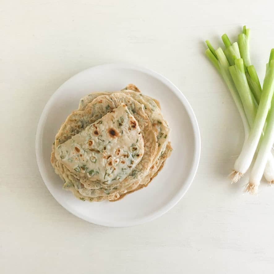 Stack of green onion pancakes on round white plate with green onions on the side