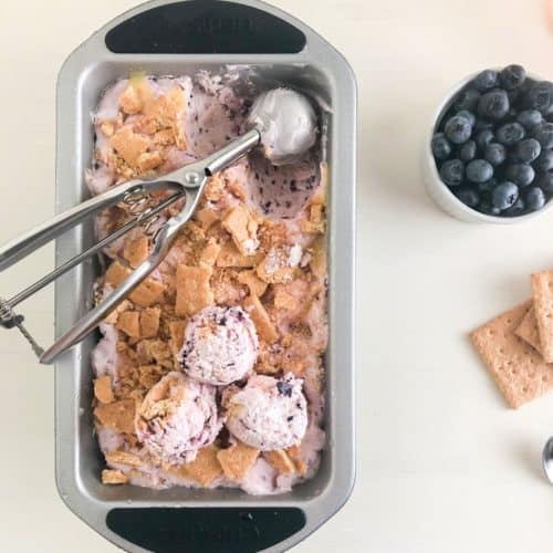 Frozen no churn ice cream in loaf pan with scoops on top and silver cookie scoop, next to small round white ramekin of blueberries and sheets of graham crackers