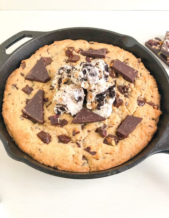 Baked cookie in black cast iron pan with scoops of ice cream on top. Chocolate bars with dried fruits and nuts in the background.