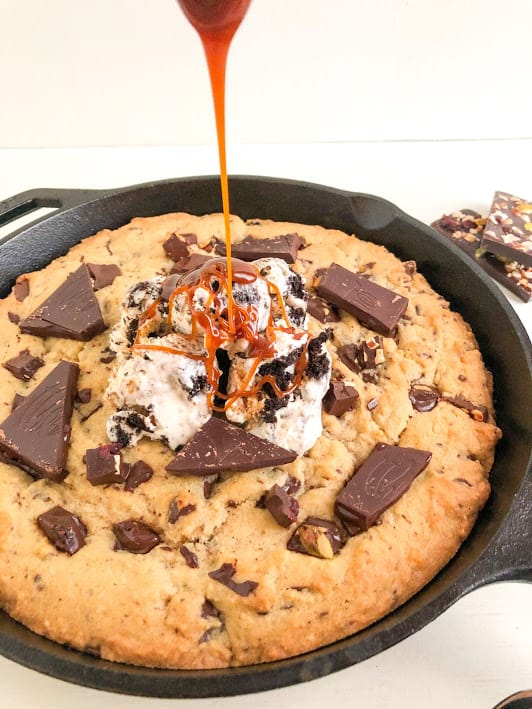 Baked cookie in black cast iron pan with scoops of ice cream, and drizzling salted caramel sauce on top of ice cream. Part of chocolate bar with dried fruit and nuts in the background.