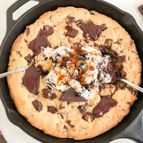 Skillet cookie in black cast iron pan with two silver spoons, scoops of ice cream, and salted caramel sauce.