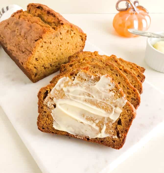 loaf of pumpkin banana bread with slices and front slice buttered on marble cutting board. Next to small white ramekin with butter and sliver butter knife, orange glass pumpkin, and white kitchen towel with gray stripes.