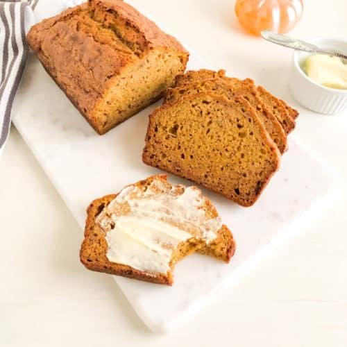 loaf of pumpkin banana bread with slices and buttered slice on marble cutting board. Next to small round white ramekin of butter with silver butter knife, with orange glass pumpkin behind. White kitchen towel with gray stripes next to loaf.