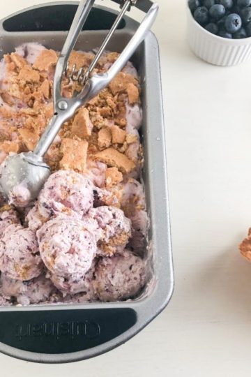 Frozen no churn ice cream in loaf pan with scoops on top and silver cookie scoop, next to small round white ramekin of blueberries, sheets of graham crackers, and waffle cones