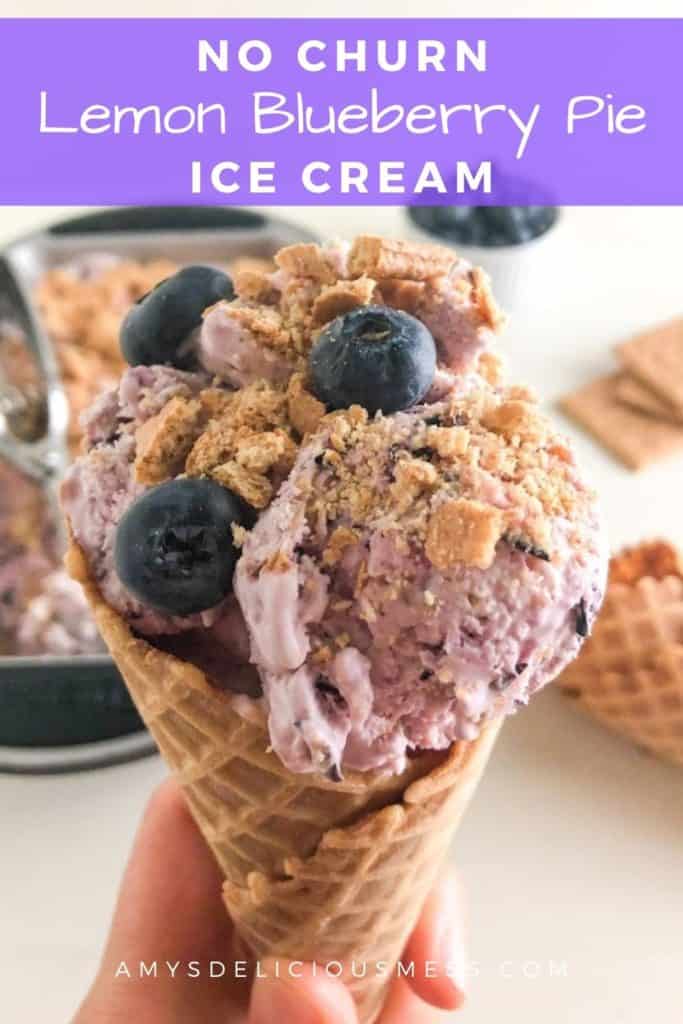 Waffle cone with ice cream, topped with fresh blueberries and graham cracker crumbs. In the background, loaf pan of ice cream with silver cookie scoop, additional waffle cones, graham cracker sheets, and white round ramekin of fresh blueberries
