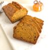 loaf of pumpkin banana bread with slices on marble cutting board. Next to orange glass pumpkin and white kitchen towel with gray stripes.